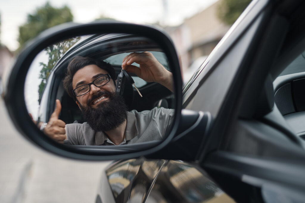 Rastreamento de carros, o cliente mais feliz. Homem carismático segurando as chaves do carro sorrindo, com dentes, enquanto está sentado no carro olhando para si mesmo no reflexo do espelho lateral.